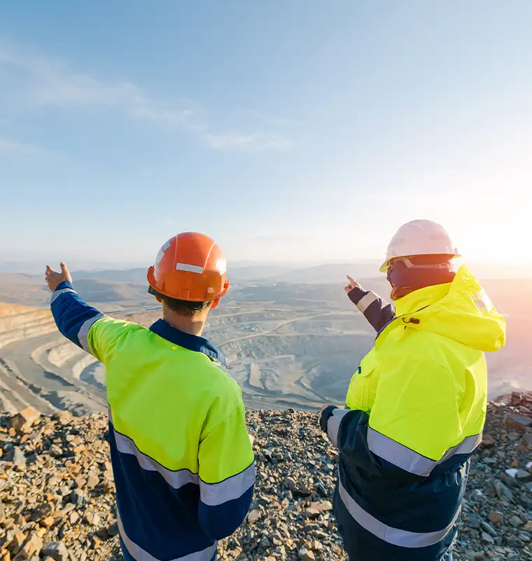 Two mineral industry professionals in hard hats and high-visibility gear surveying an open-pit iron ore mine representing UMI's global sourcing expertise