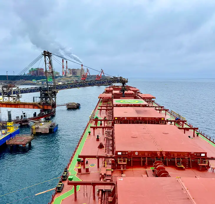 Deck view of a red-hulled bulk carrier at an industrial port terminal with loading cranes representing UMI's global iron ore shipping and logistics services